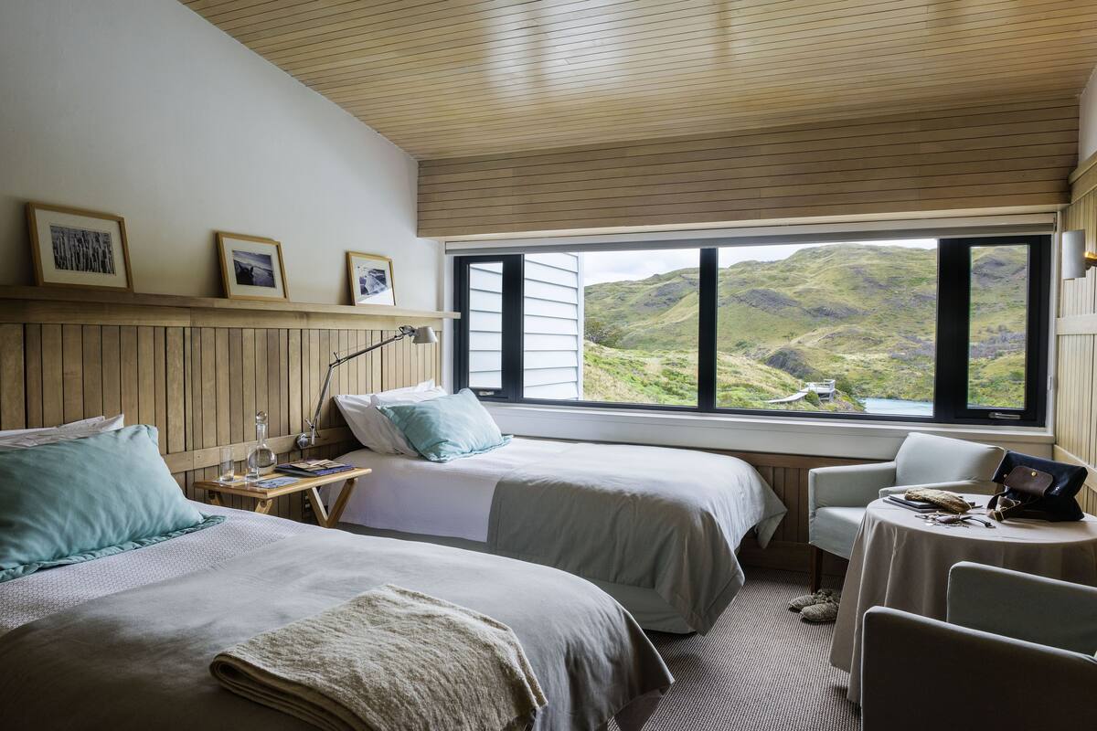 A bedroom with two beds, a desk, and a chair, with a view of a mountainous landscape through a large window.