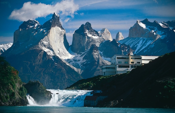 A modern hotel with a mountain backdrop, a waterfall, and a clear sky.