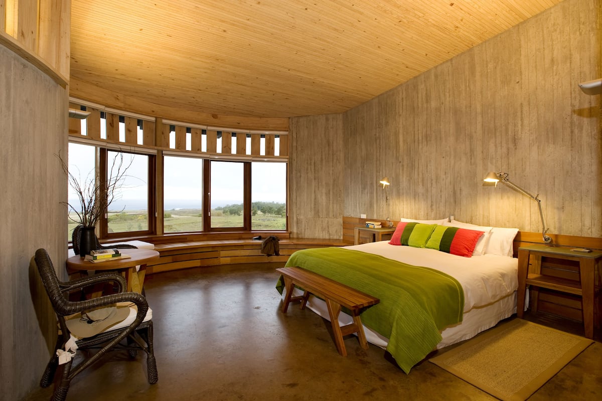 A bedroom with a large bed, wooden headboard, a bench, a chair, a desk, and a window with a view of the outdoors.