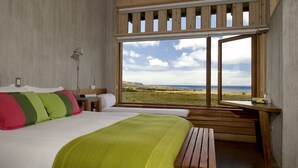 A bedroom with a large window, a bed with green and pink pillows, a bedside table, and a wooden bench. - explora Rapa Nui (Hanga Roa)