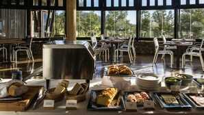 A buffet with various bread, pastries, and a coffee station in a spacious room with large windows and a stone wall. - explora Rapa Nui (Hanga Roa)
