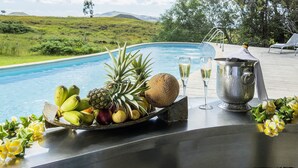 A poolside table with fruits, glasses, and a bucket, overlooking a pool with a scenic view of hills and trees. - explora Rapa Nui (Hanga Roa)
