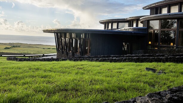 A modern building with a curved roof and large windows, situated on a grassy field with a view of the ocean. - explora Rapa Nui (Hanga Roa)