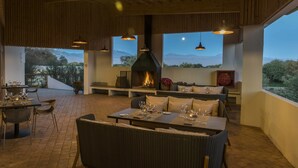 Un área de comedor al aire libre con mobiliario de mimbre, una chimenea y grandes ventanales que ofrecen vistas a la montaña.