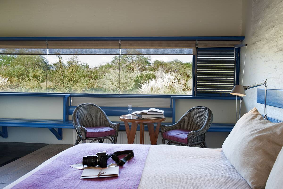 A bedroom with a bed, two wicker chairs, a small table, and a window with blinds.