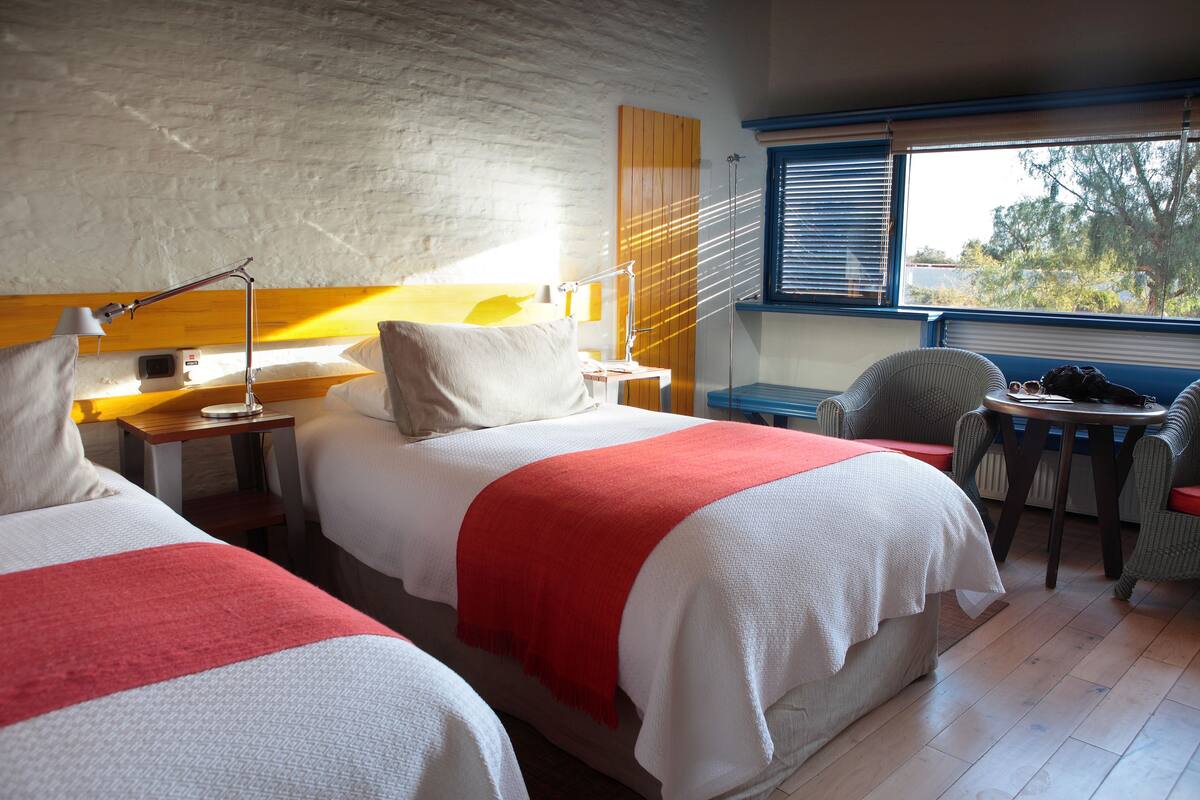 A hotel room with two beds, a small table, a chair, a window with blinds, and a view of trees outside.