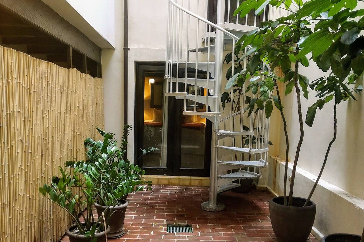 Ein Innenhof mit Backsteinpflasterung, Topfpflanzen, einer Wendeltreppe und einer Bambuswand.