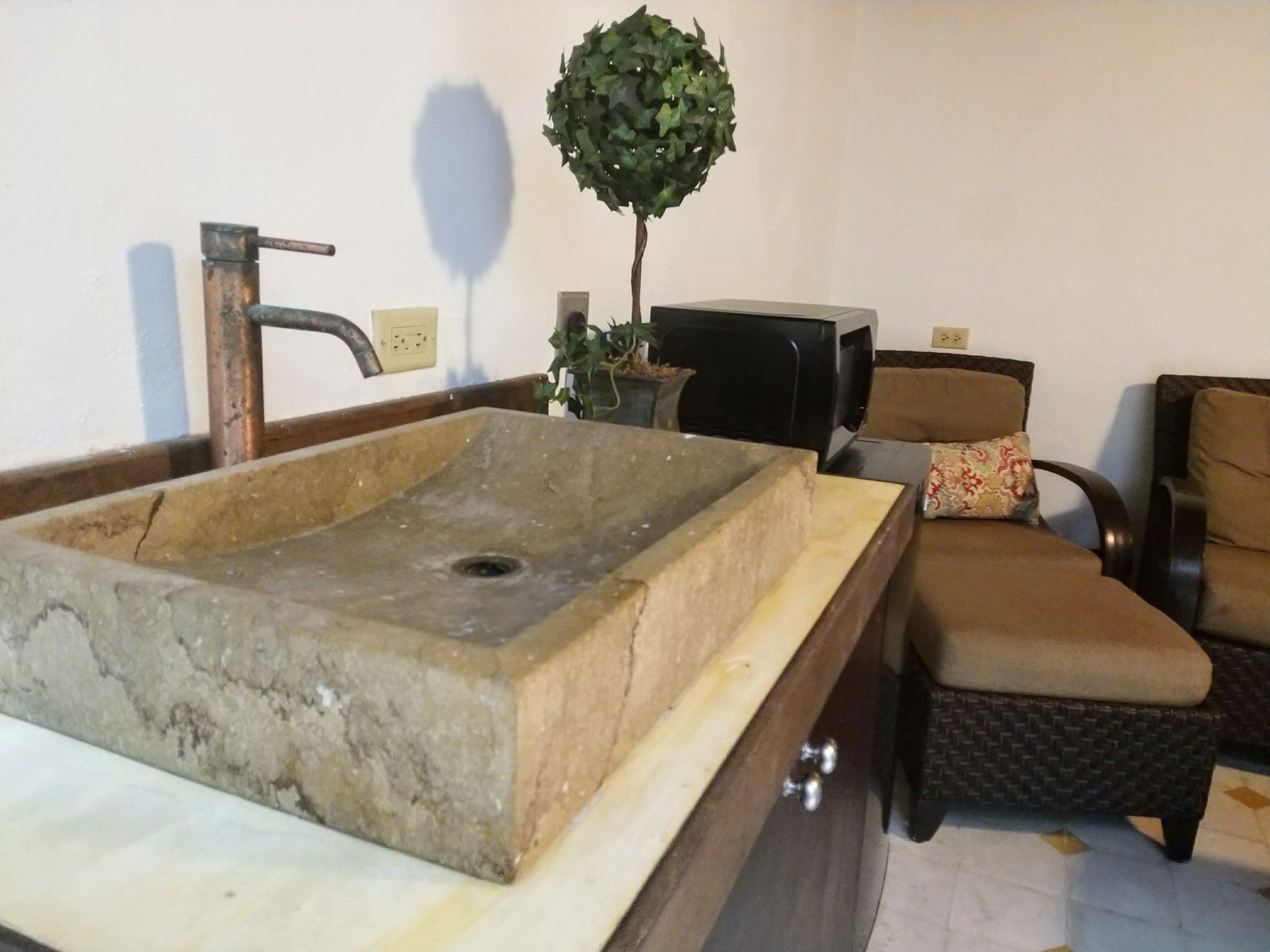 A bathroom with a rectangular stone sink, a faucet, a potted plant, a television, and a brown armchair.