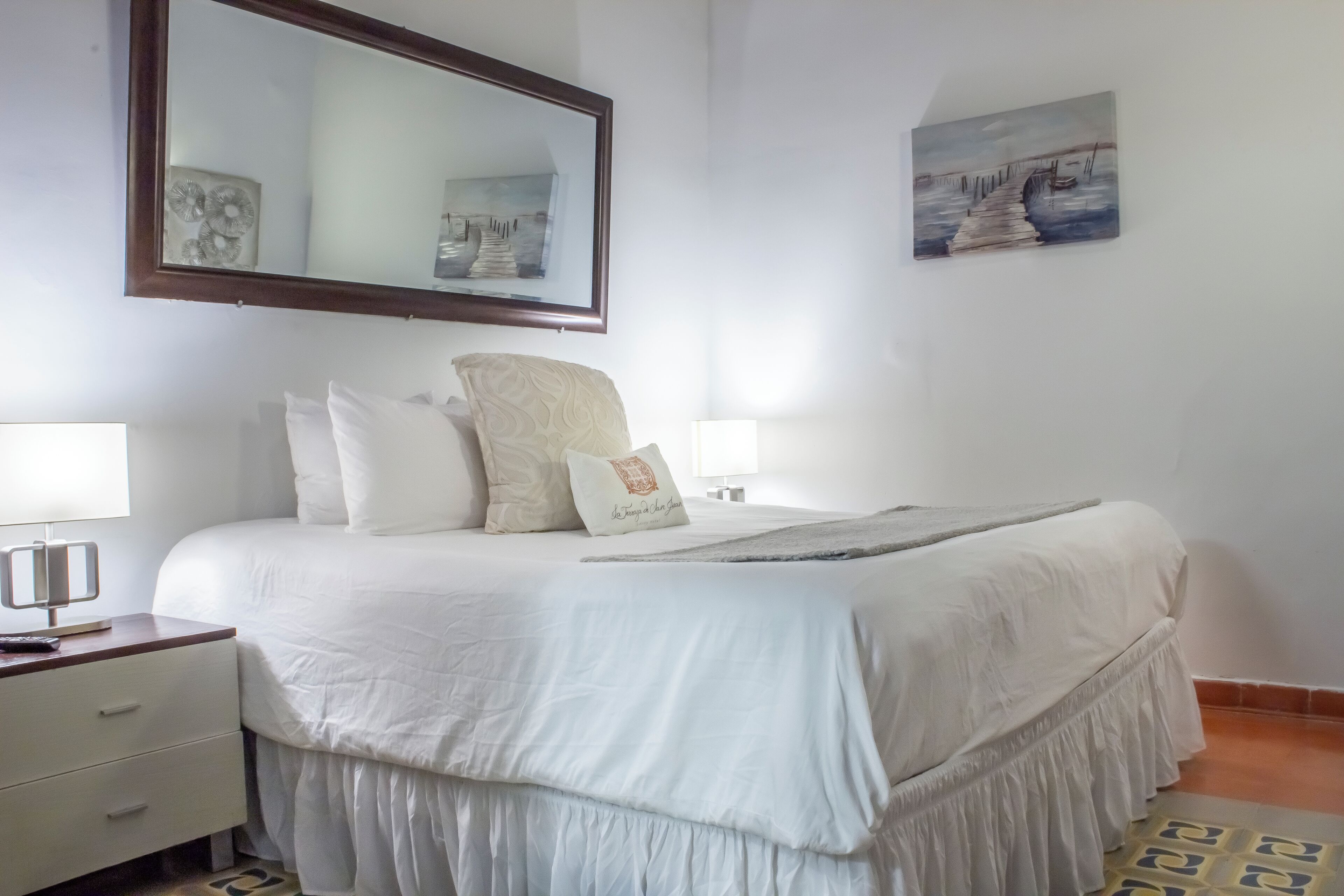 Una cama bien hecha con ropa blanca, una lámpara de noche y un cojín decorativo.