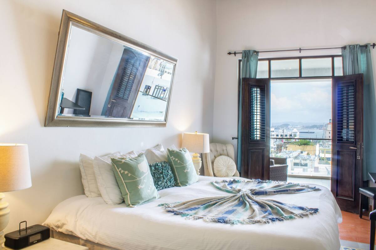 Ein Hotelzimmer mit einem großen Bett, Blick auf die Stadt durch das Fenster und einem Spiegel an der Wand.