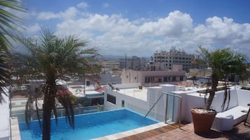 A rooftop pool with palm trees, a clear blue sky, and city buildings in the background.