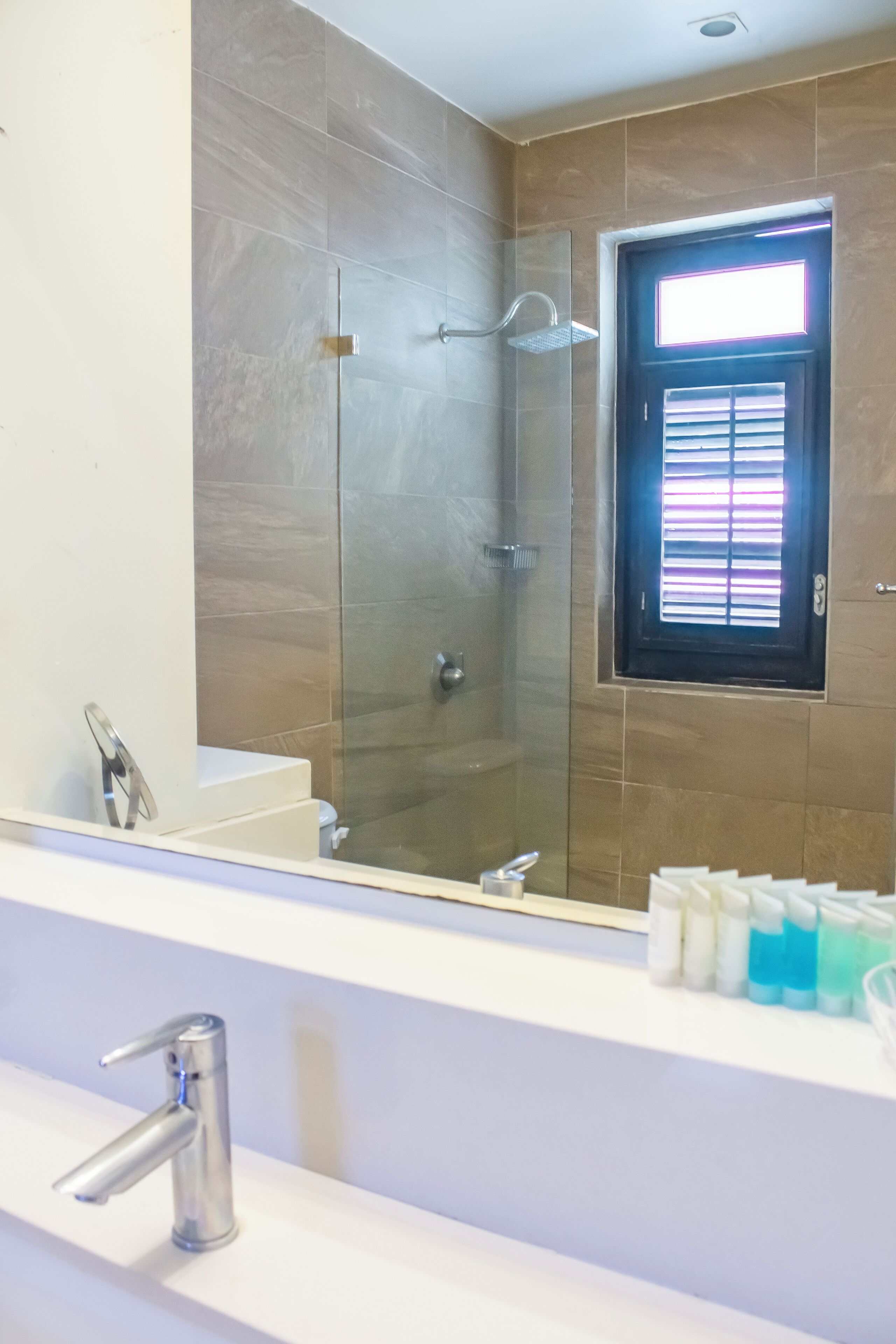 Un baño con ducha envuelta en vidrio, un lavamanos con grifo moderno y una ventana con persianas.