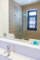 Un baño con ducha envuelta en vidrio, un lavamanos con grifo moderno y una ventana con persianas.