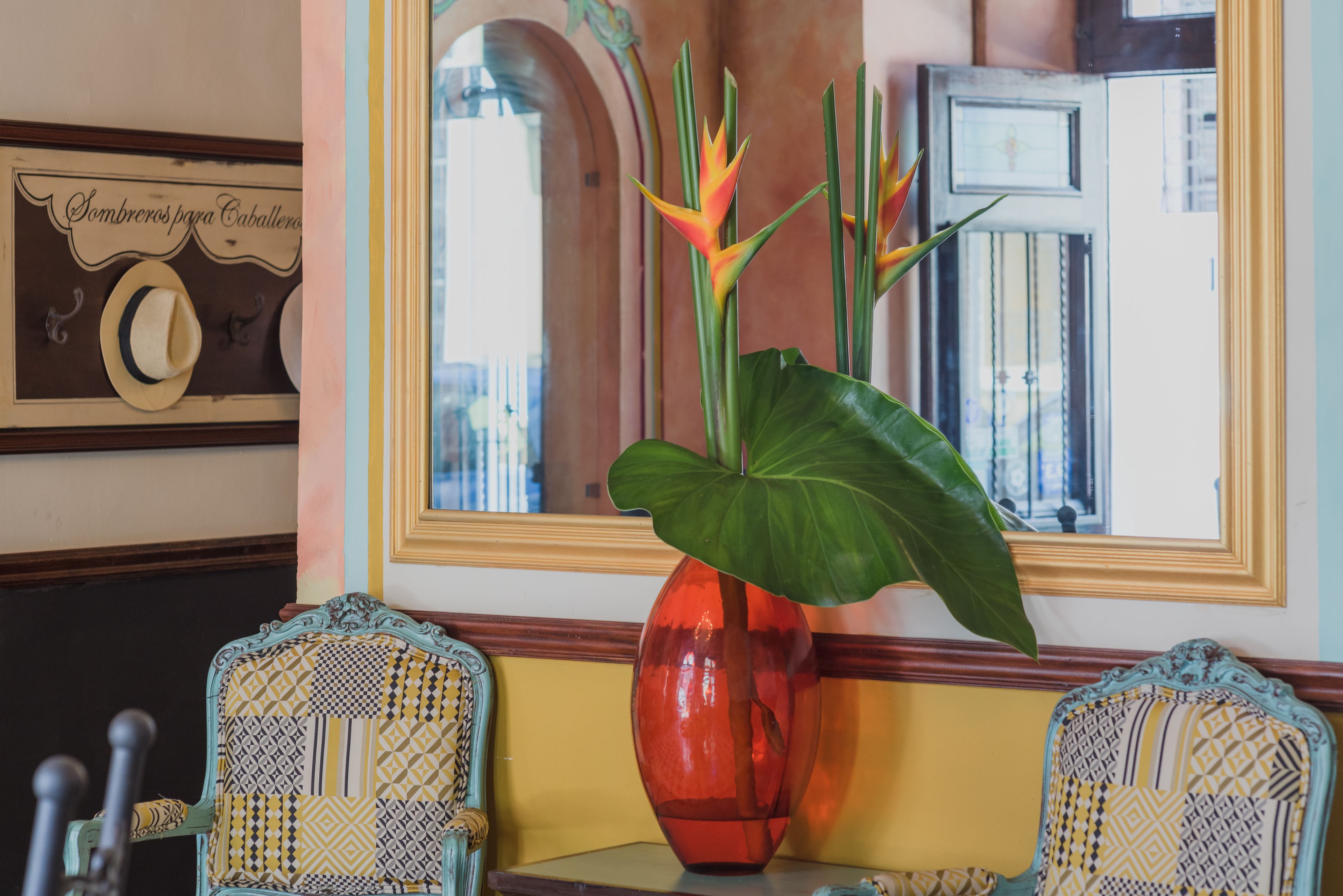 Una habitación con un espejo grande, un florero rojo con una planta y sillas estampadas.