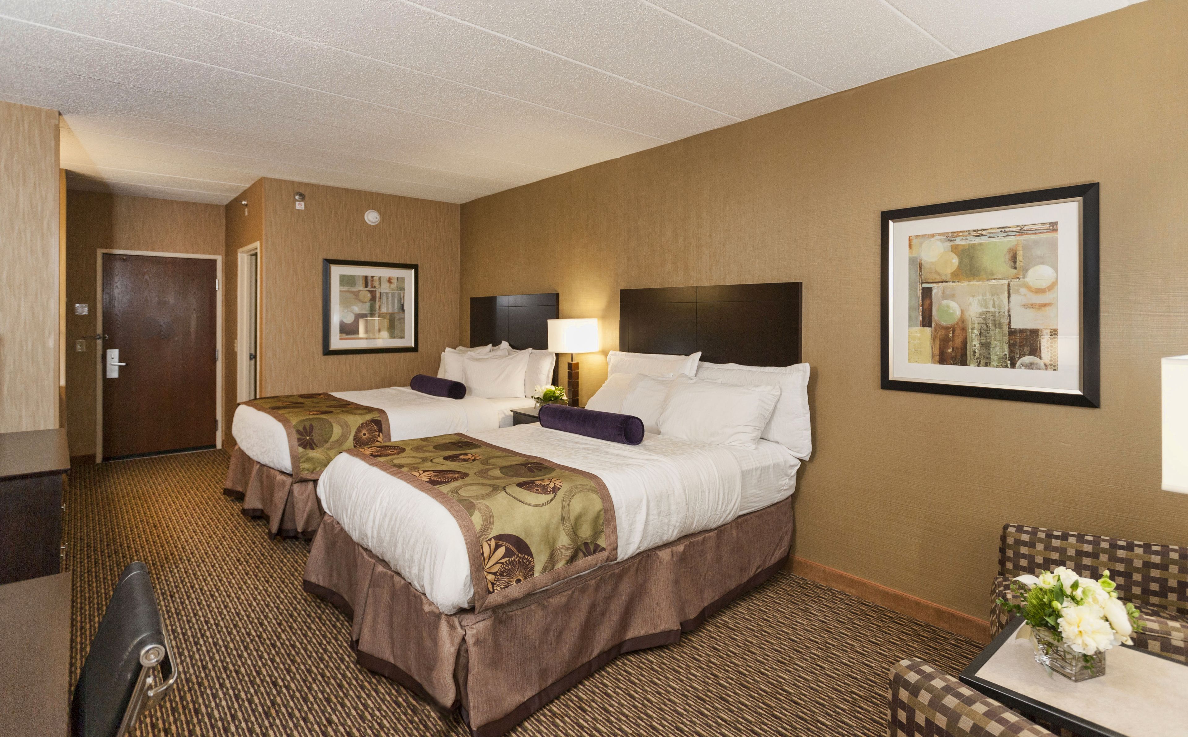 A hotel room with two beds, a desk, a chair, a lamp, and framed pictures on the walls.