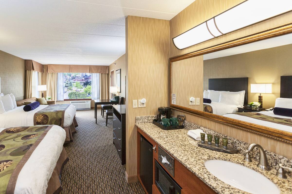 A hotel room with two beds, a desk, a large mirror, a sink, and a view of the outdoors.