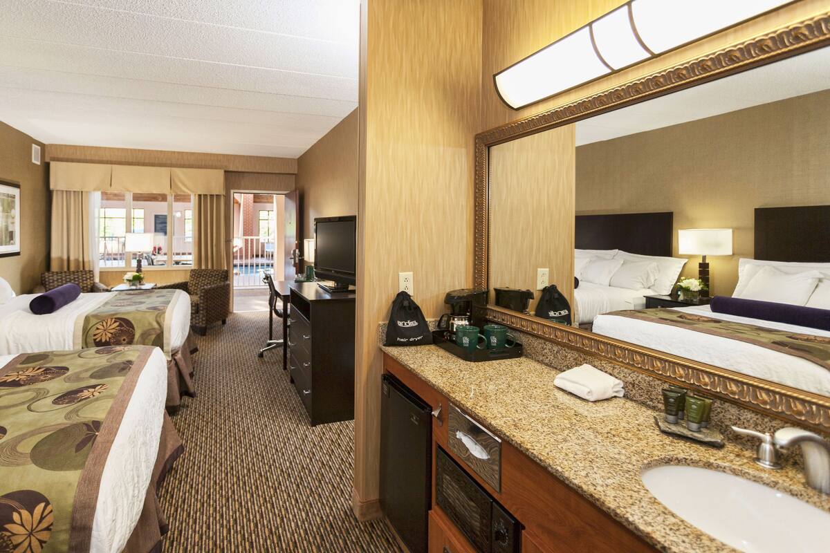 A hotel room with two beds, a desk, a television, a coffee maker, and a bathroom with a sink and mirror.
