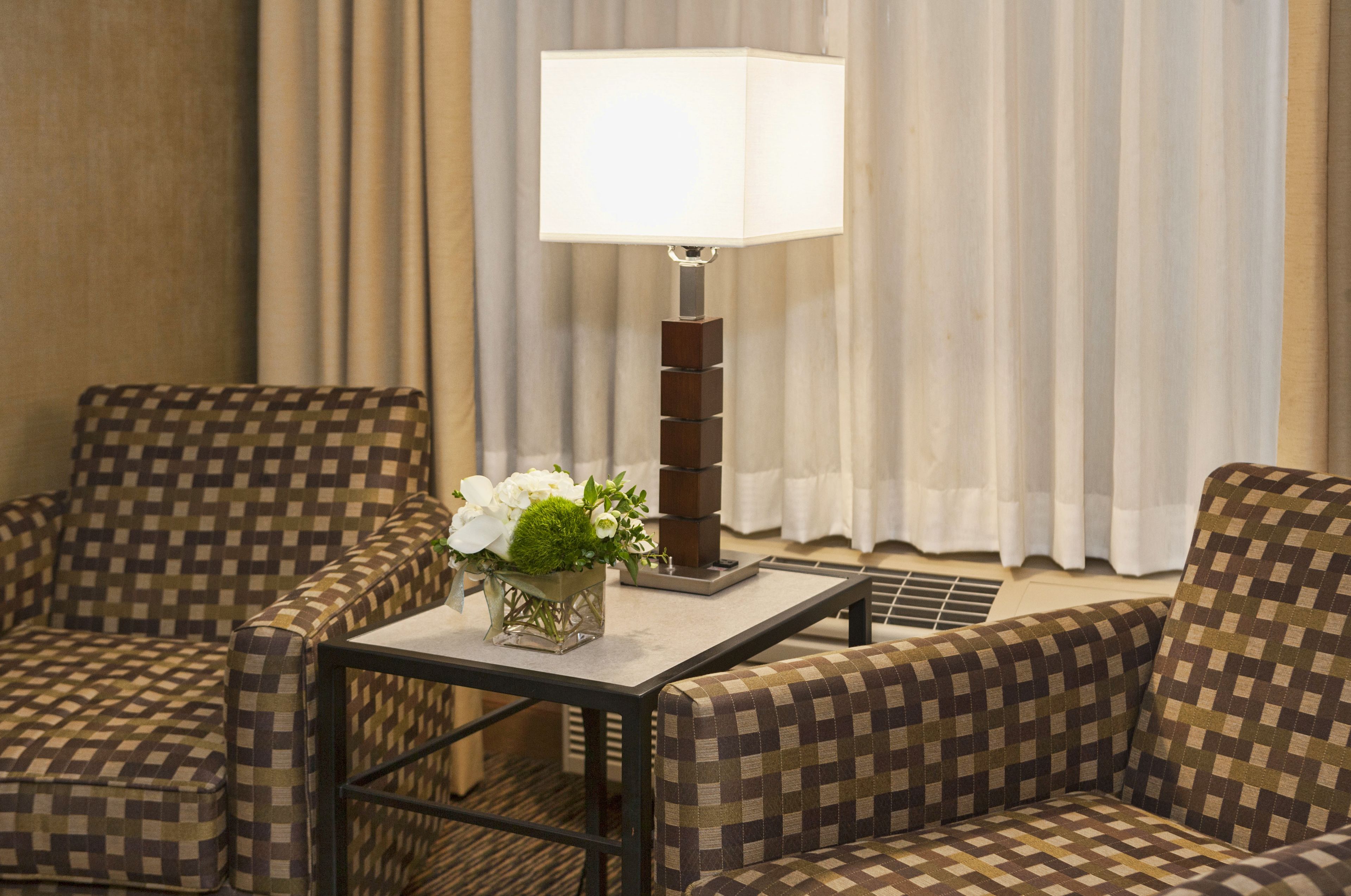 A hotel room with checkered armchairs, a table with a lamp, and a vase with flowers.