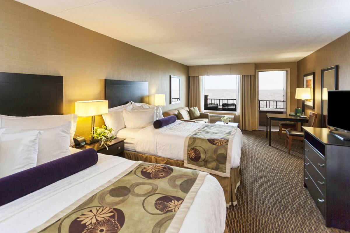 A hotel room with two beds, a desk, a chair, a TV, and a balcony with a view of the sea.