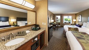 Une chambre d’hôtel avec un grand miroir, un lavabo, un four à micro-ondes et un bureau avec une chaise. La chambre est équipée de deux lits avec des oreillers et d’une fenêtre donnant sur un paysage verdoyant.