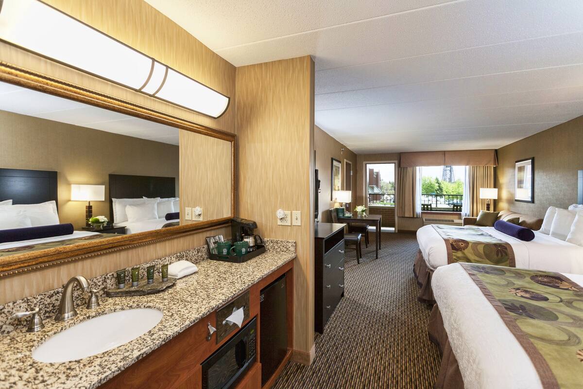 A hotel room with a large mirror, a sink, a microwave, and a desk with a chair. There are two beds with pillows and a window with a view of greenery.