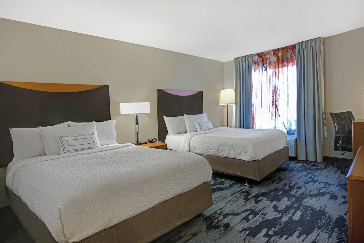 Quarto de hotel com duas camas, uma escrivaninha, uma cadeira e uma janela grande com cortinas.
