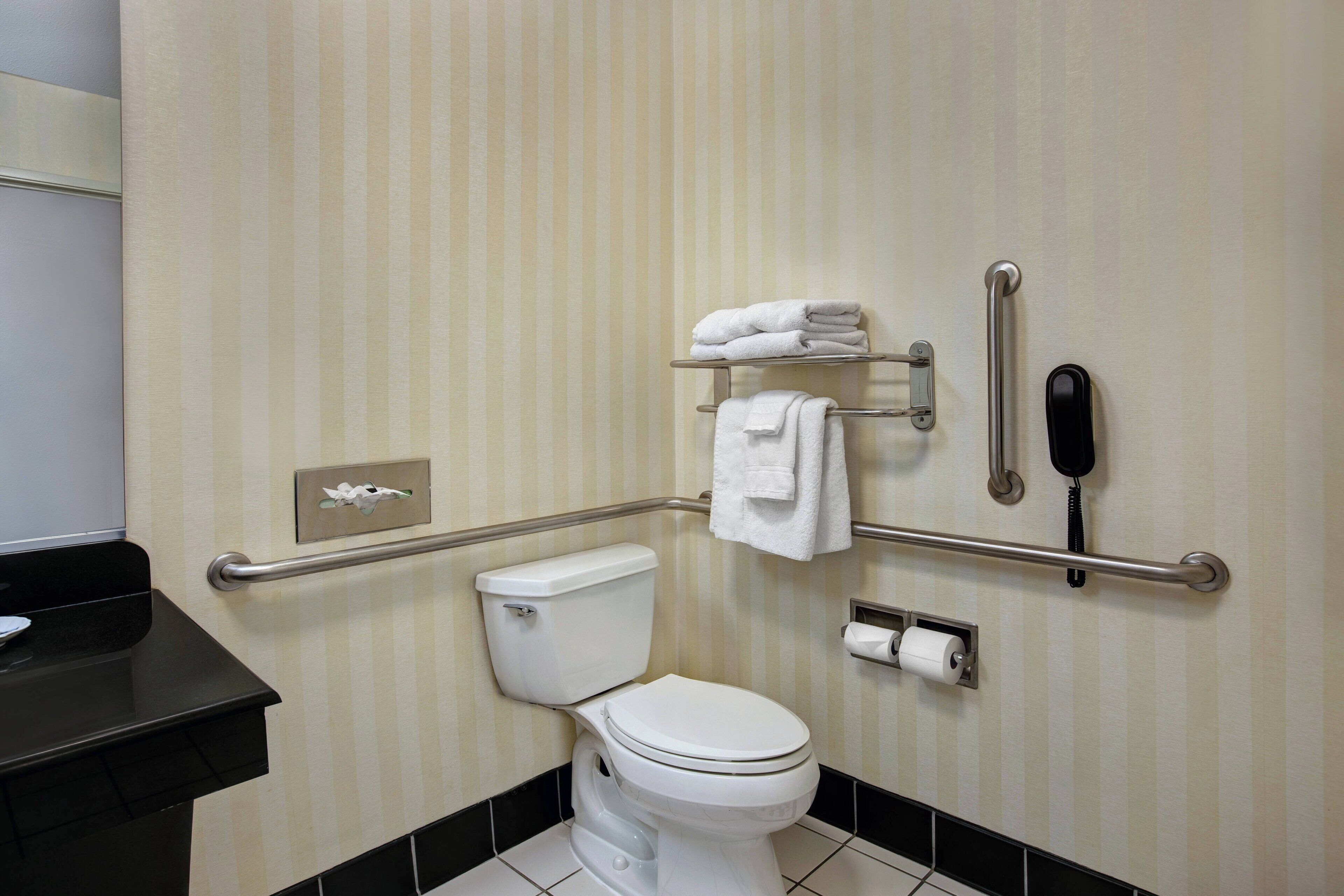 Et badeværelse med toilet, gelændere, et håndklædestativ, en telefon og en toiletpapirholder.