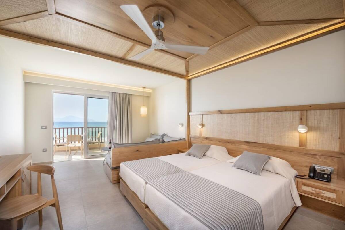 A hotel room with a bed, bedside table, and a desk with chairs. There is a balcony with a view of the sea.
