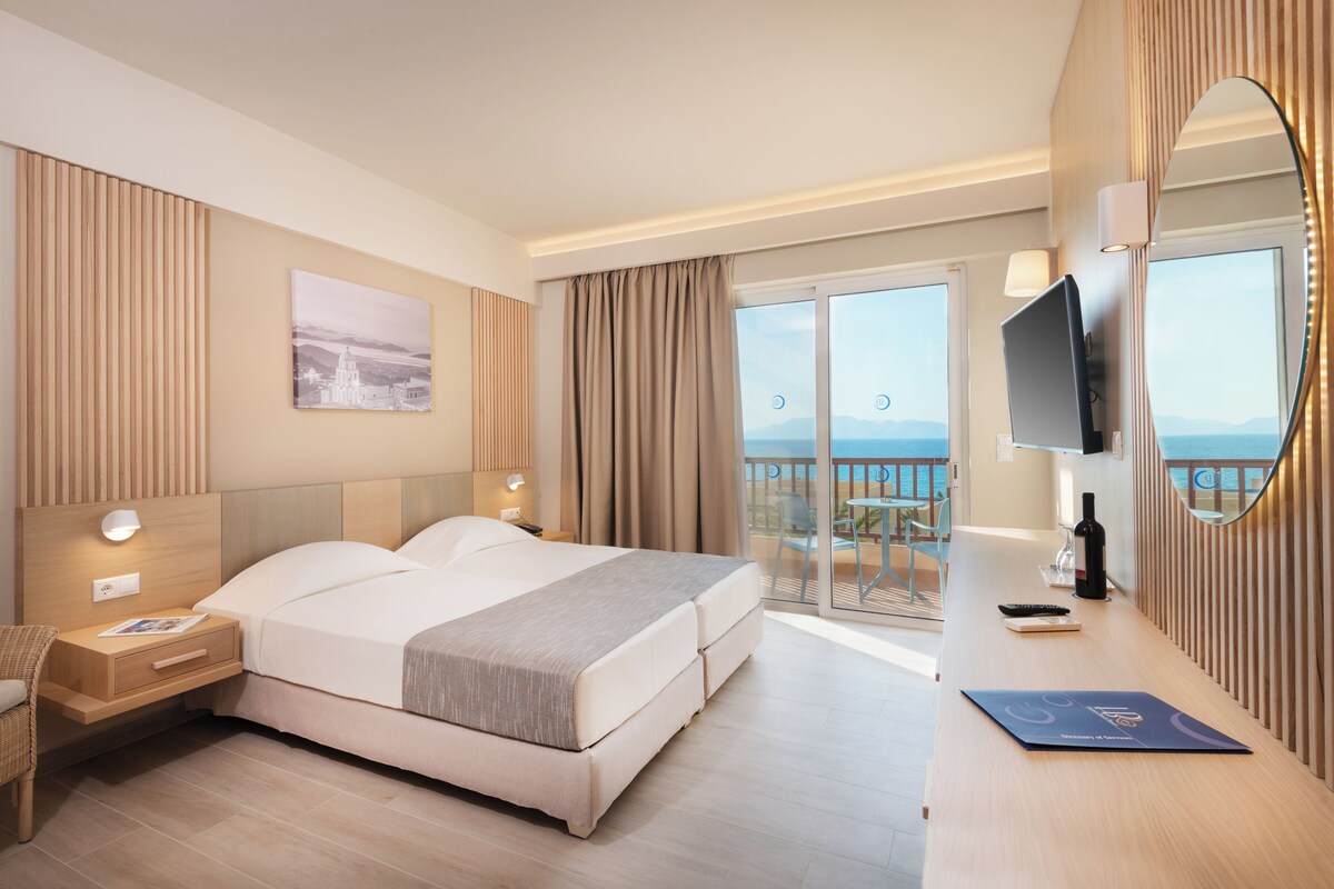 A hotel room with a bed, bedside tables, a desk with a laptop, a TV, and a balcony with a view of the sea.