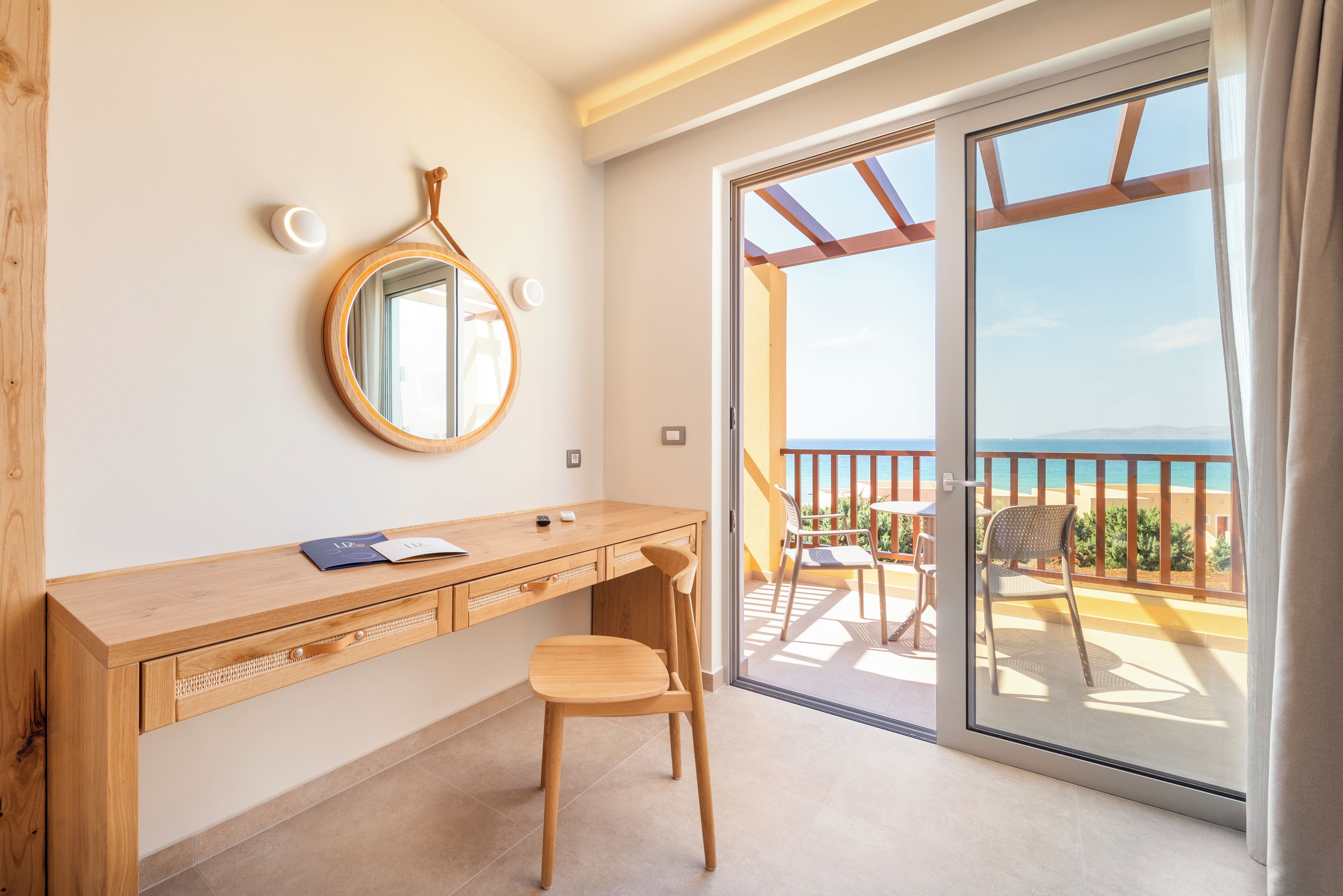 A room with a wooden desk, a chair, a round mirror, and a sliding glass door leading to an outdoor balcony with a view of the sea.