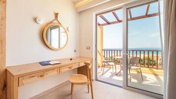 A room with a wooden desk, a chair, a round mirror, and a sliding glass door leading to an outdoor balcony with a view of the sea.