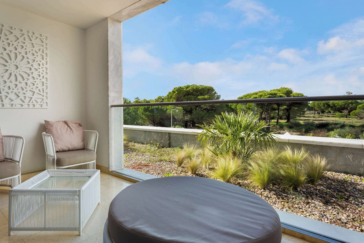 A balcony with a view of a landscaped garden, a white chair, a sofa, and a glass coffee table.