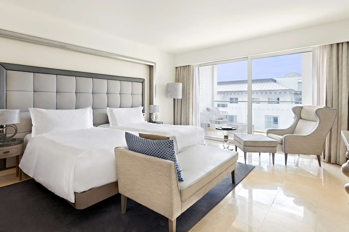 A hotel room with a large bed, two bedside tables with lamps, a sofa, a chair, and a balcony with a view of buildings.