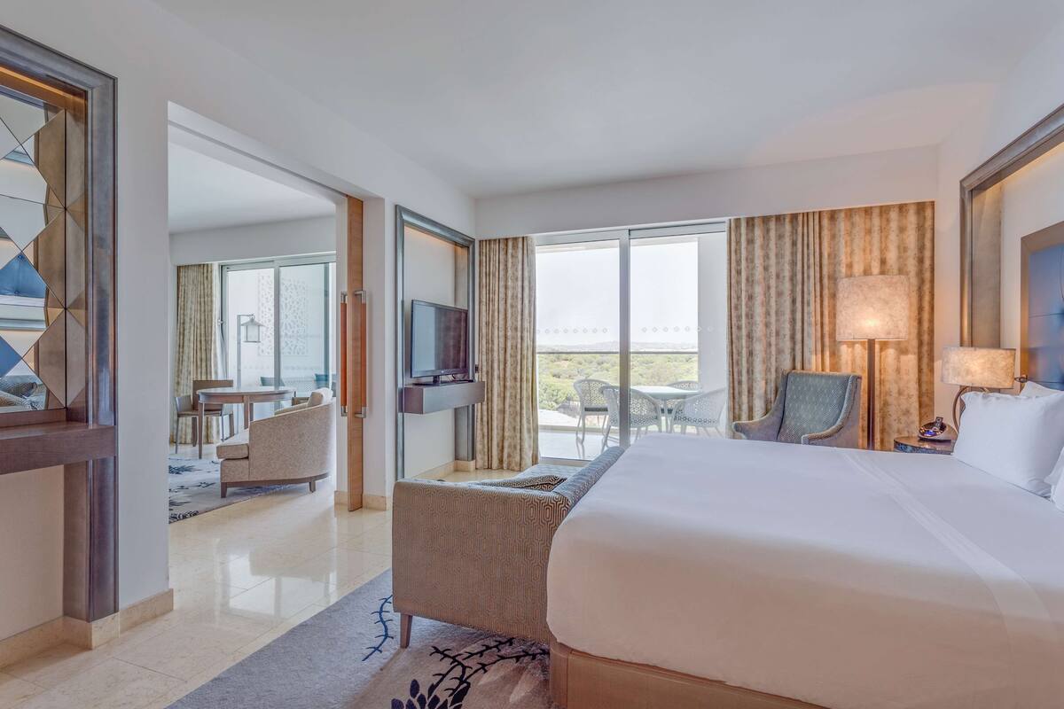A hotel room with a large bed, a TV mounted on the wall, a seating area with a table and chairs, and a balcony with a view.