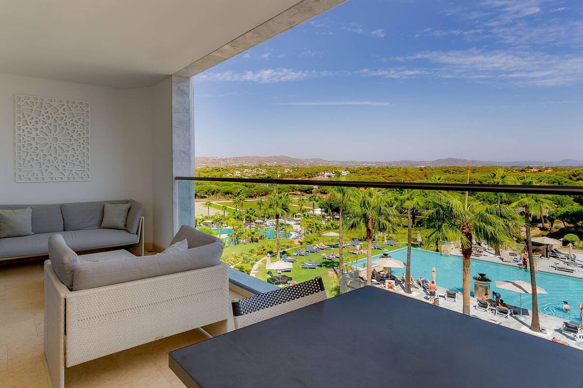 A balcony with a sofa, a wicker chair, and a view of a pool and resort area.