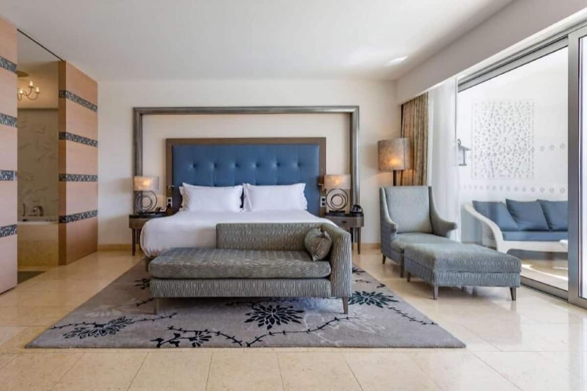 A modern hotel room with a large bed, a sofa, a chair, and a large window.