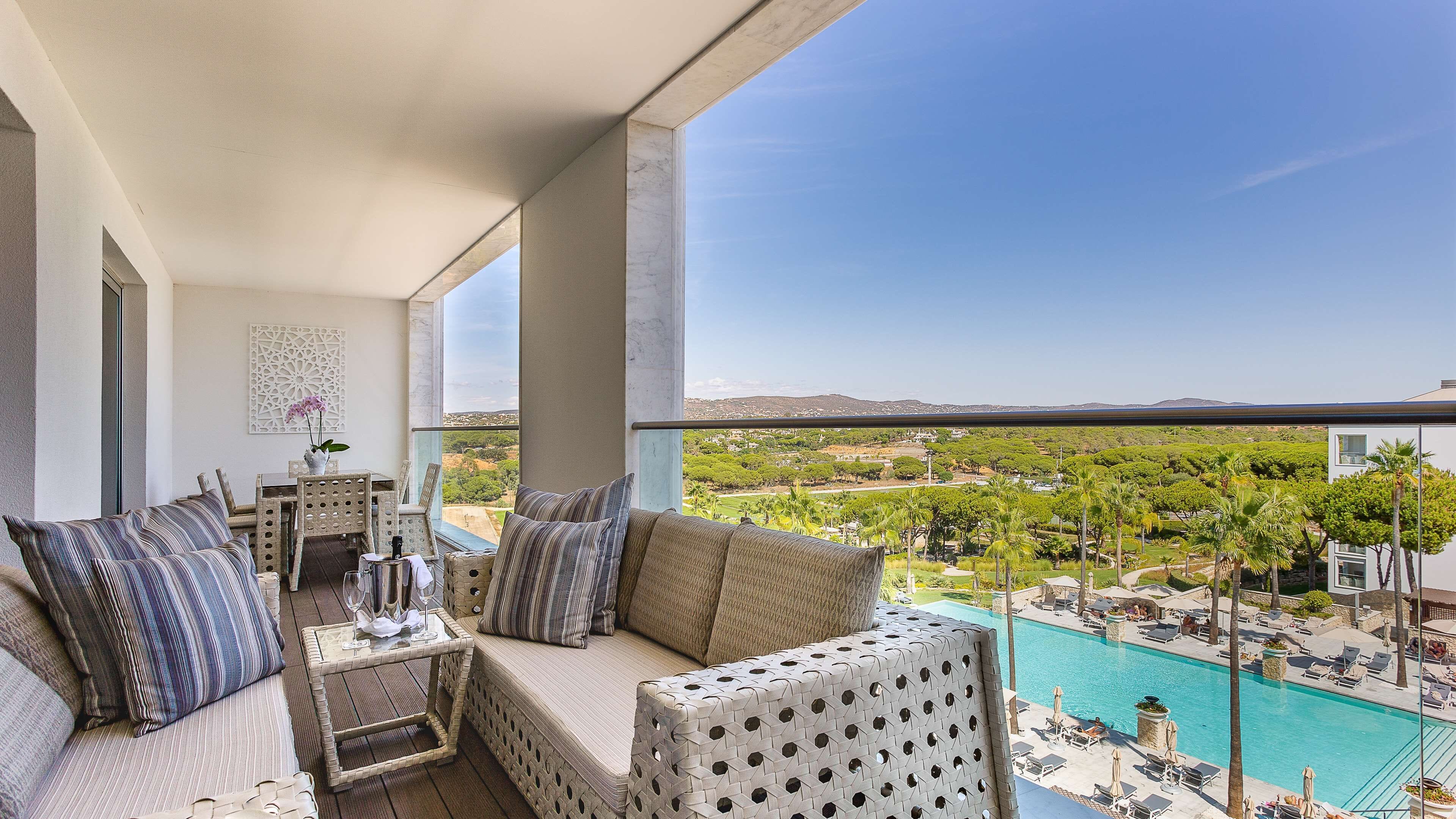 A balcony with a sofa, dining table, and chairs, overlooking a swimming pool and lush greenery.