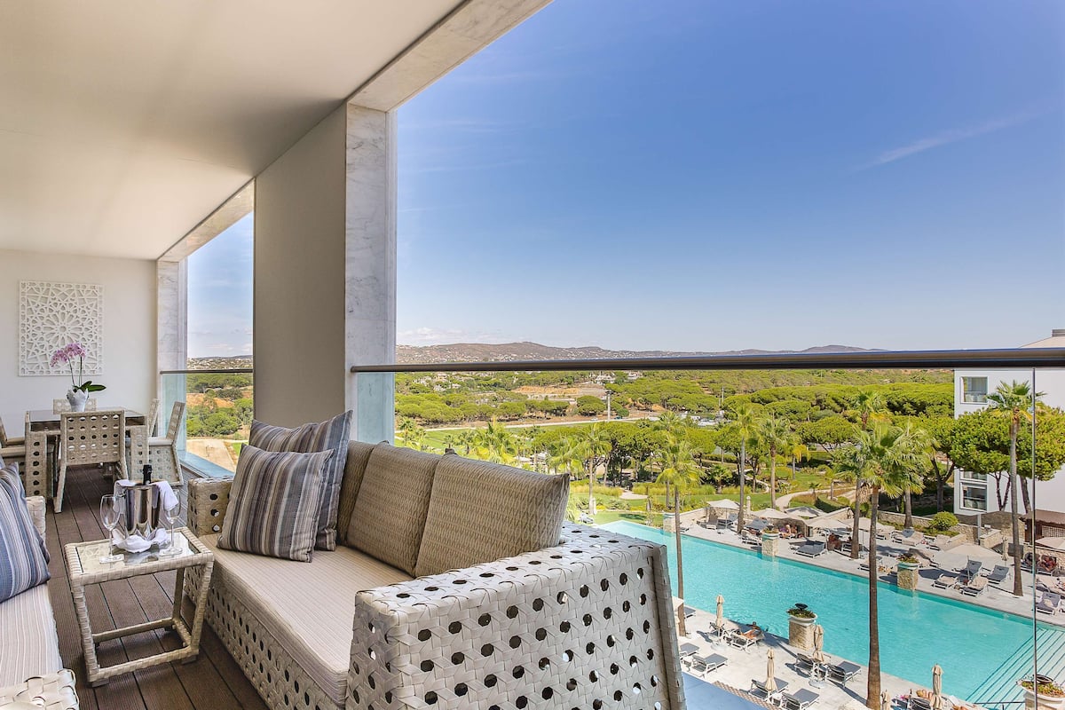 A balcony with a sofa, dining table, and chairs, overlooking a swimming pool and lush greenery.