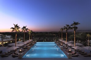 Zona de piscina con sillas de descanso y sombrillas, rodeada de palmeras, al caer la noche.