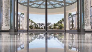 A modern hotel lobby with a glass entrance, palm trees, and a reflective floor.