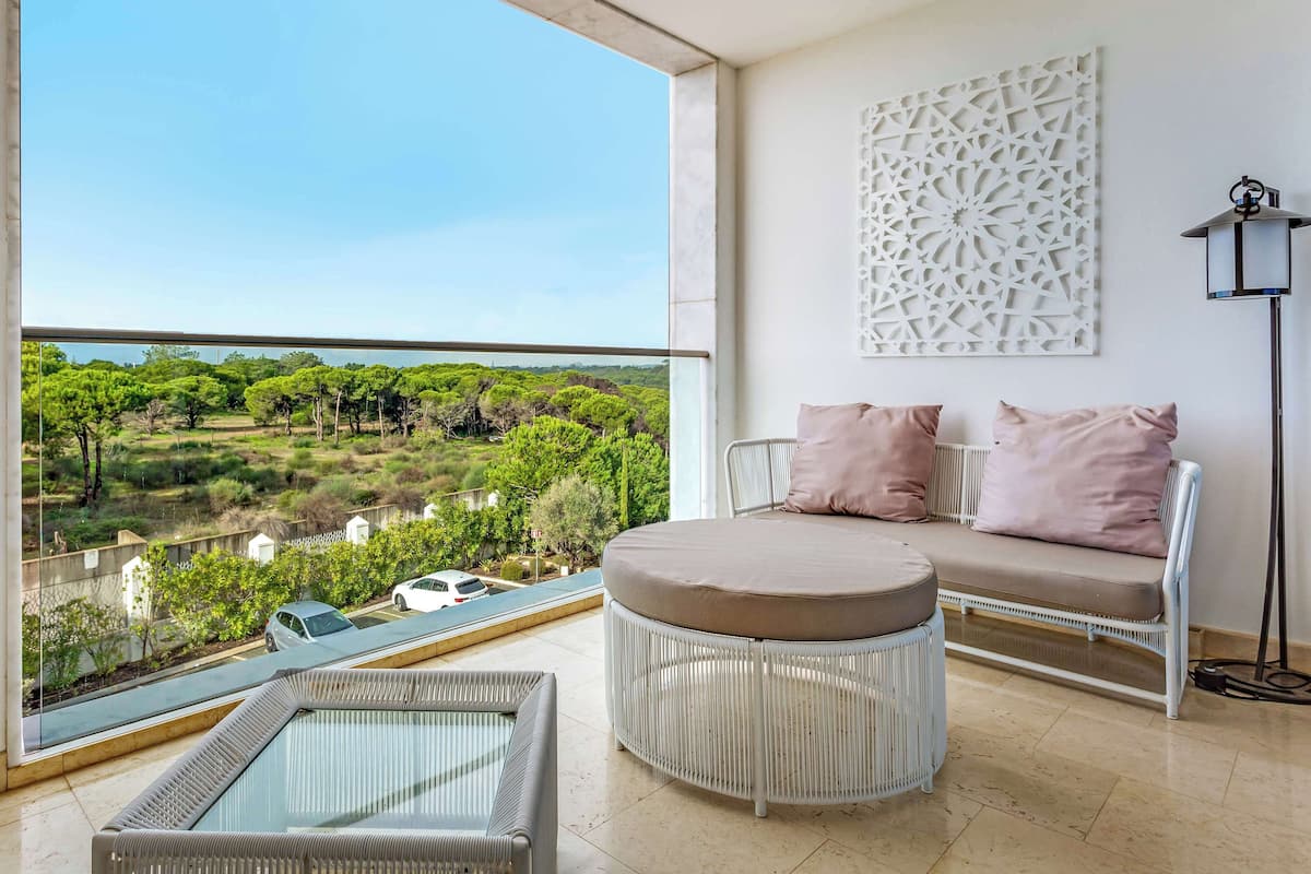 A balcony with a view of a green landscape, a white sofa with cushions, a round wicker chair, and a floor lamp.