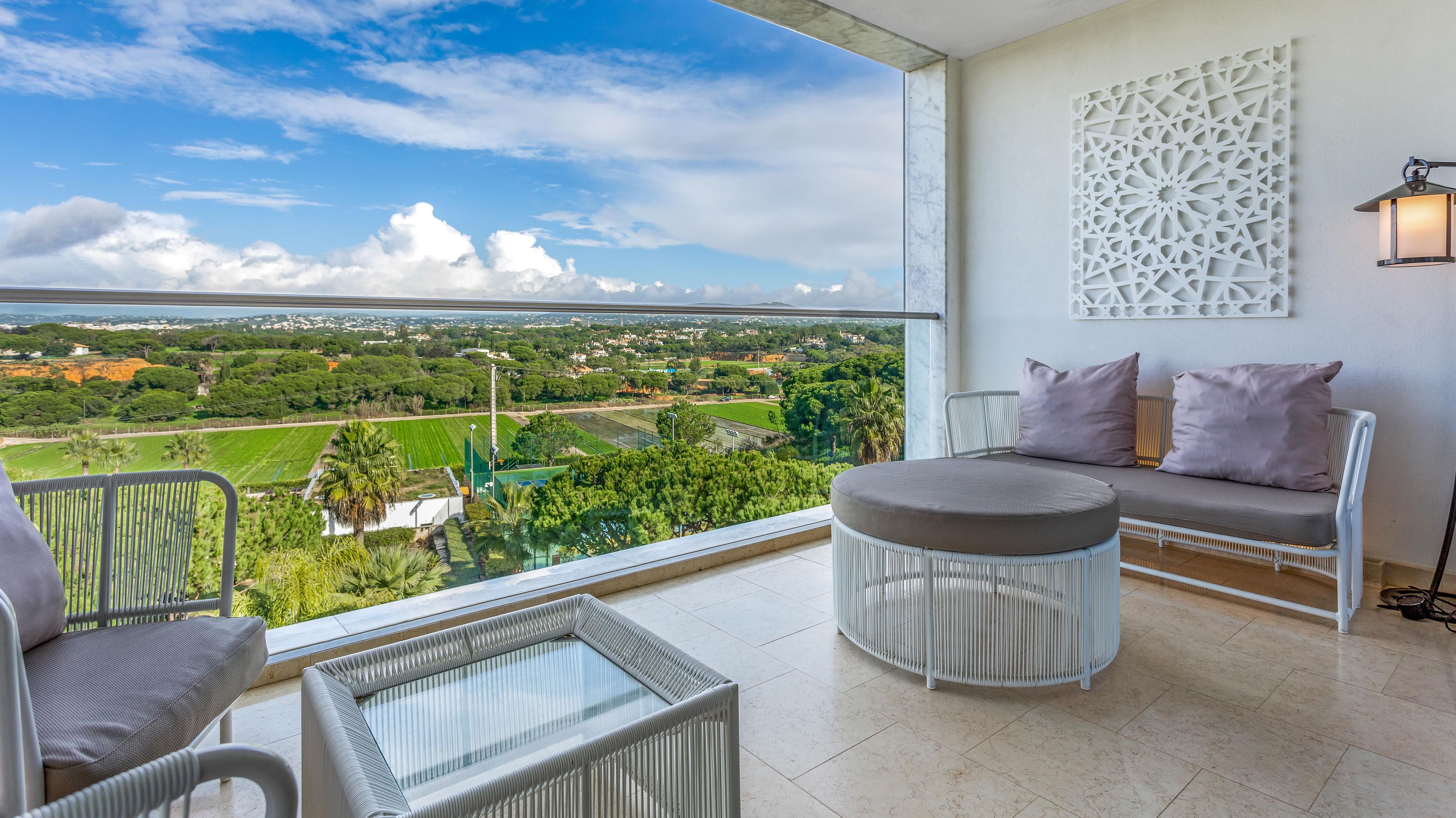 Un balcón con vista a un paisaje verde, una silla blanca, un reposapiés redondo y una lámpara fijada a la pared.