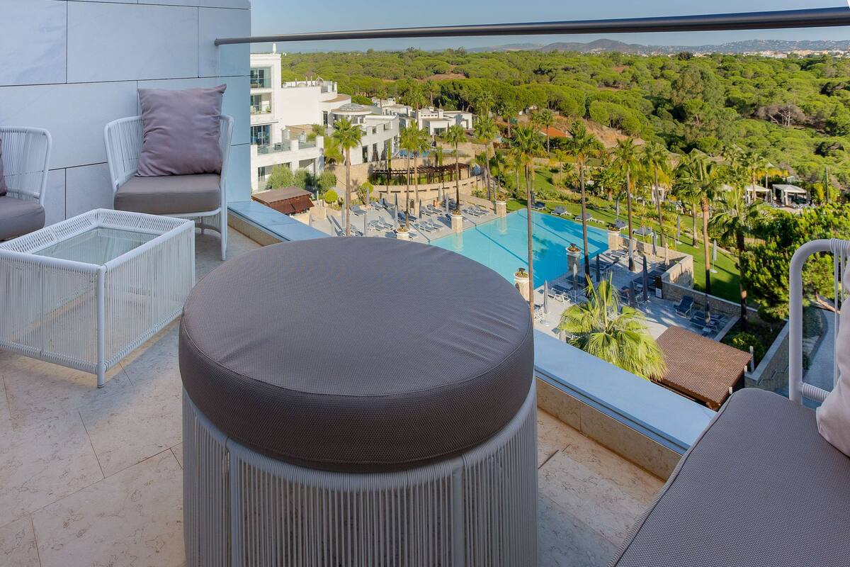A balcony with wicker furniture, a glass coffee table, and a view of a pool and greenery.
