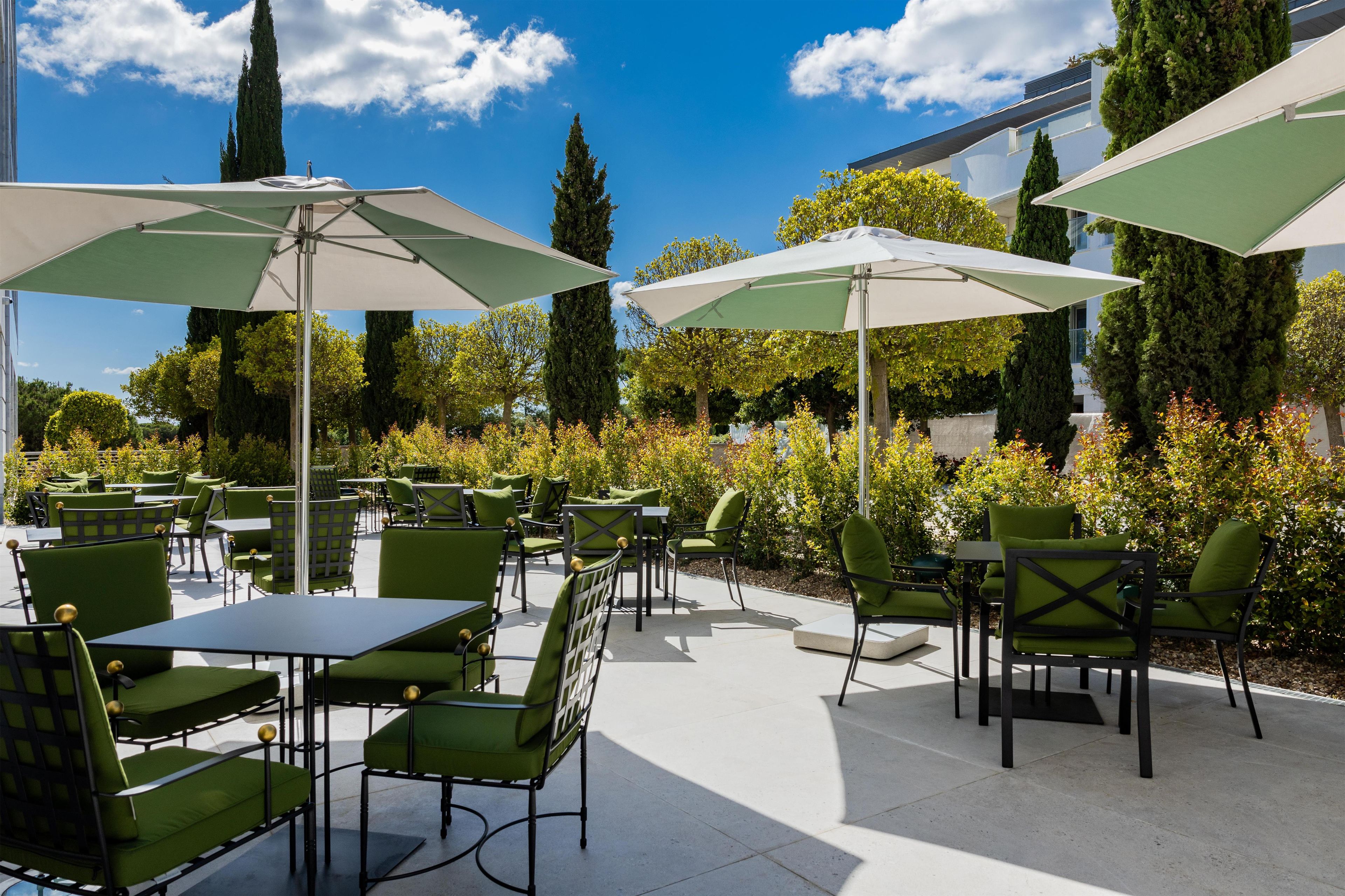 An outdoor patio with green chairs and tables, umbrellas, and a backdrop of trees and shrubs.
