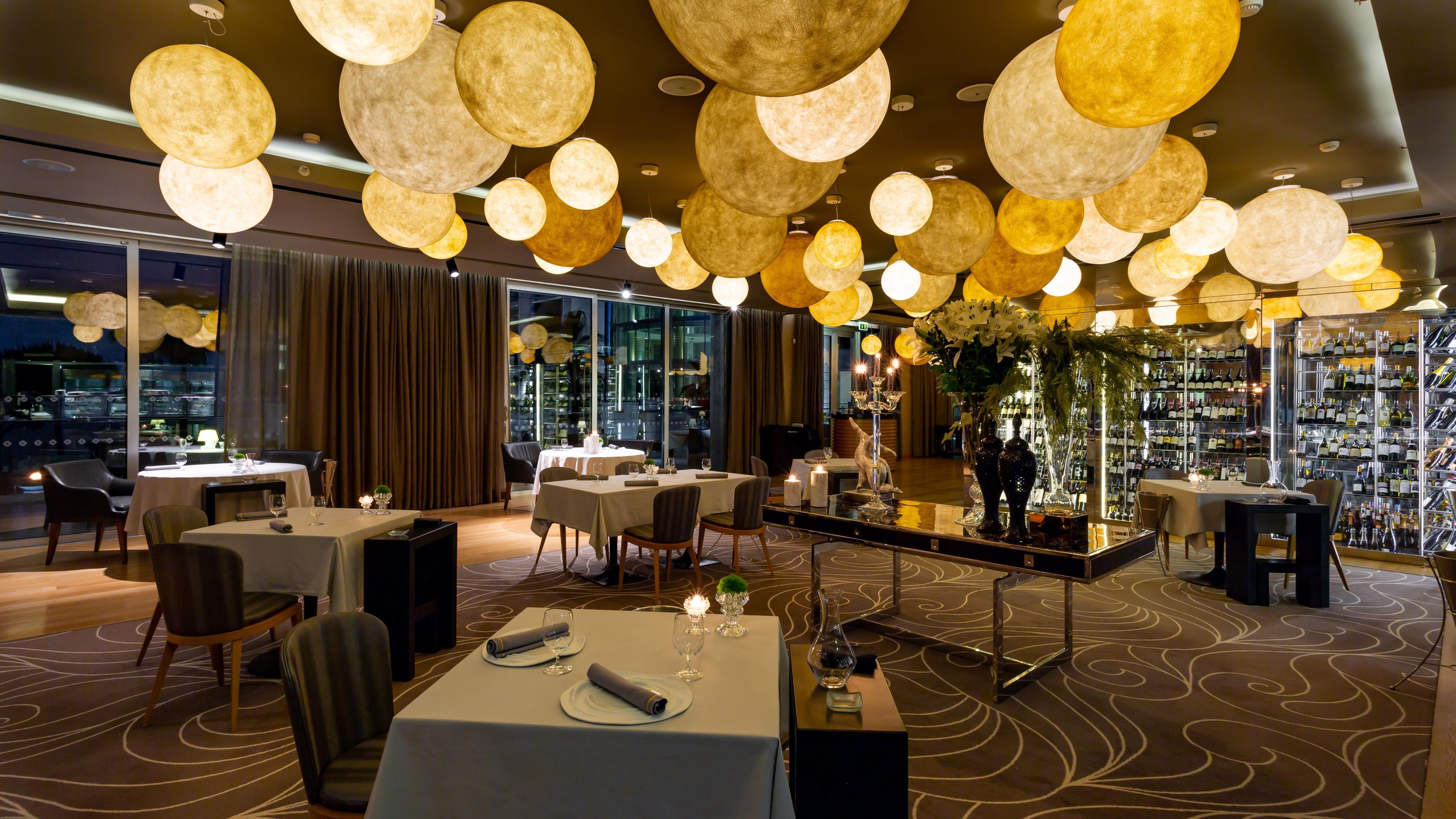 A restaurant with round, illuminated hanging lights, tables set for dining, and a wine display.