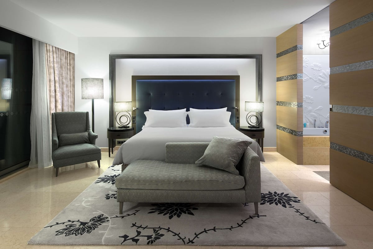 A modern hotel room with a large bed, a sofa, a chair, two bedside tables with lamps, and a bathroom visible through a sliding door.