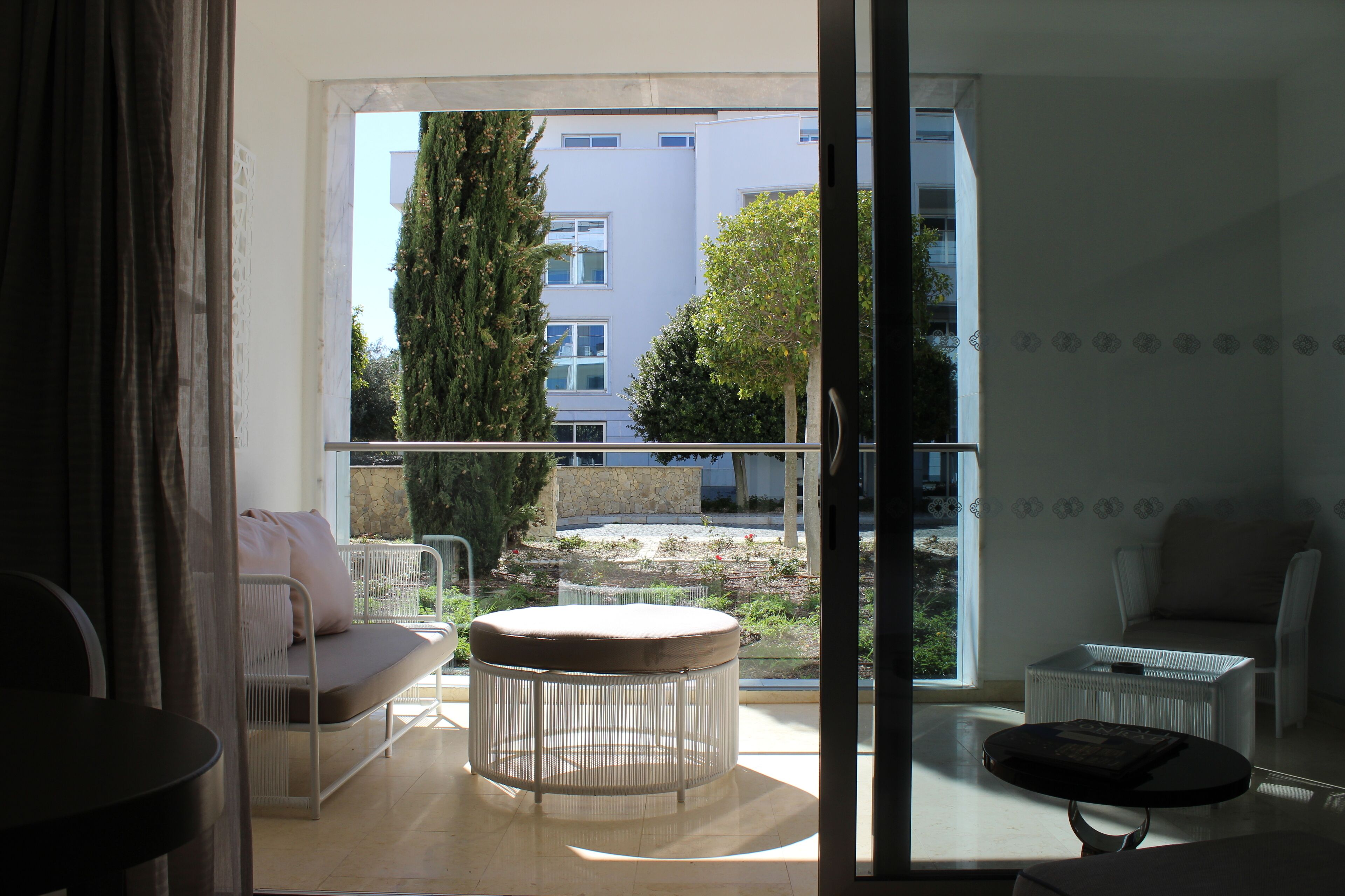 A modern living room with a sofa, a round ottoman, and a small table. A large window provides a view of a building and greenery outside.