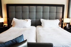 Habitación de hotel con dos camas, cada una con ropa de cama blanca y un cabecero oscuro. Hay dos mesitas de noche con lámparas y un reloj pequeño en cada una.