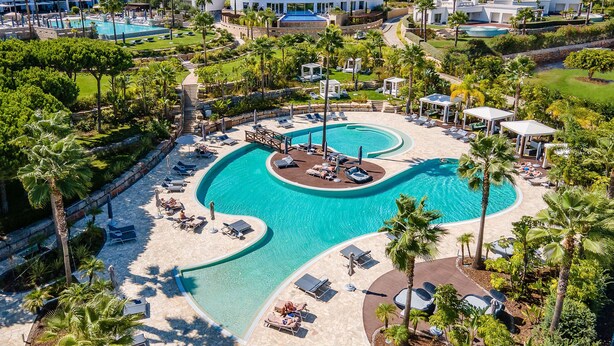 A resort with a large central pool, sun loungers, and palm trees.