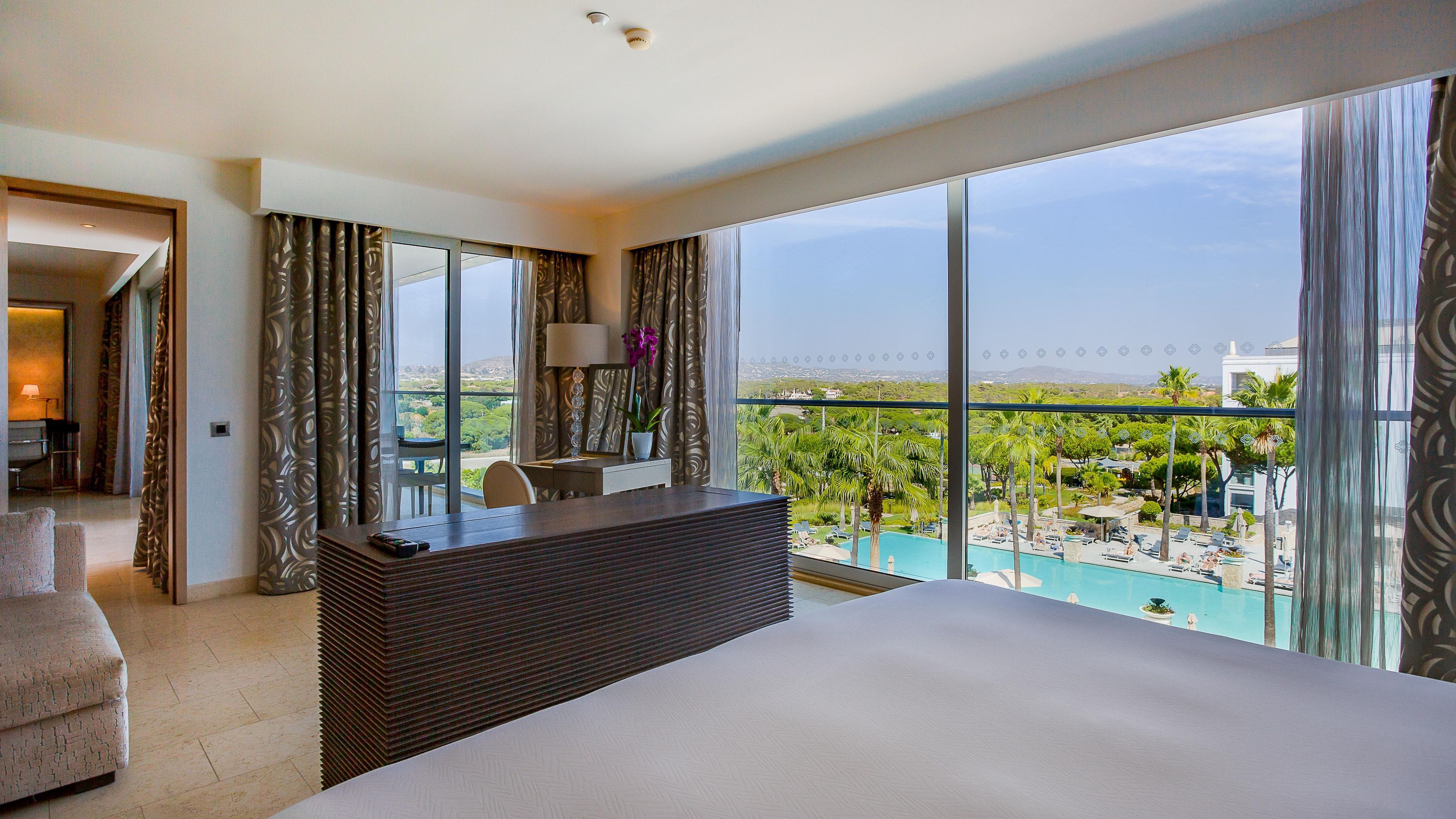 A hotel room with a large bed, a desk, a chair, and a view of a swimming pool and greenery.